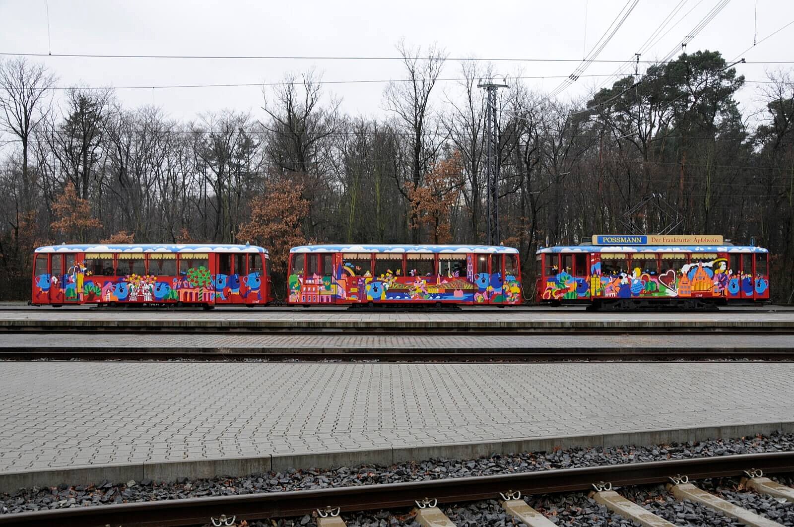 Drei Ebbelwei-Express Wagons hintereinander auf den Gleisen vor Bäumen.